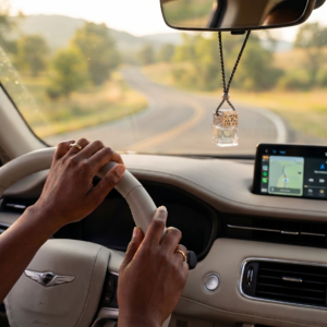 A serene, first-person view from the driver's seat of a sophisticated vehicle cabin. The hands of an African American woman, adorned with elegant gold rings, rest comfortably on the cream leather steering wheel. Hanging from the rearview mirror is a clear glass car diffuser with a silver lattice lid. The car is traveling along a quiet, winding road at sunset, with soft, golden light filling the cabin and evoking a feeling of total peace and clarity.