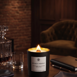 A premium matte black candle with a glowing gold interior and a lit wood wick, centered on a dark wood table in a sophisticated, moody lounge setting with a leather chair and crystal decanter in the background.