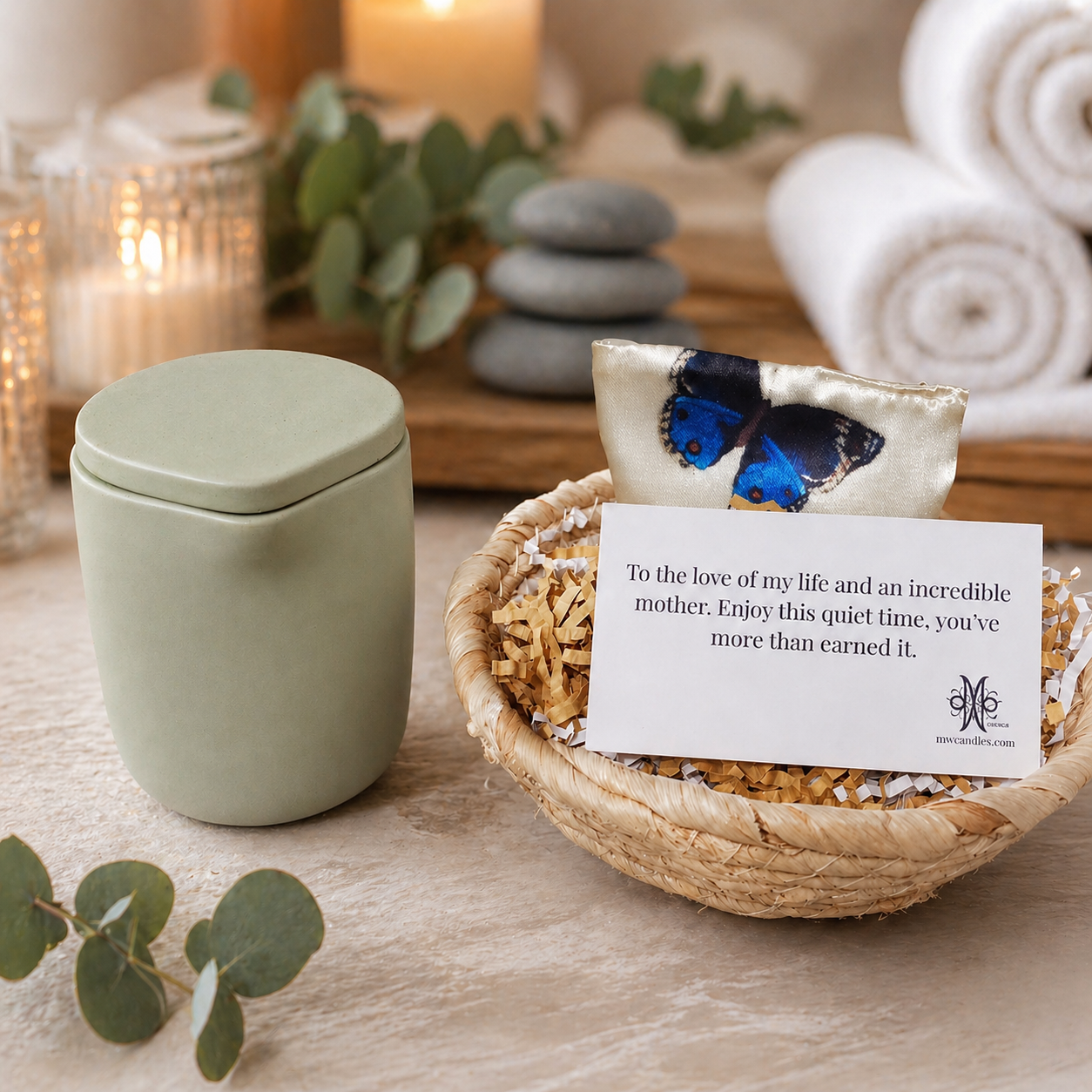 A mint-green, spouted concrete botanical massage candle sits on a natural wood surface next to a small woven basket. Tucked inside the basket is a soft, satin sleep eye mask with a butterfly pattern. The background is a serene, spa-inspired setting featuring two rolled white towels, fresh eucalyptus leaves, and smooth gray stones. A few glass candles are lit in the soft-focus background, providing a peaceful, ambient glow.